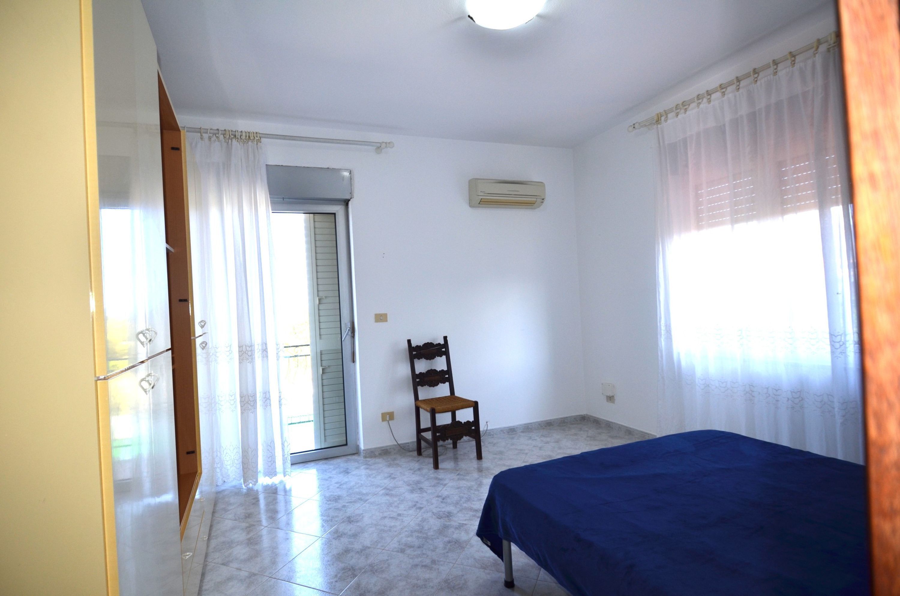 A room with a bed, a chair, a window with curtains, an air conditioning unit, and a door.