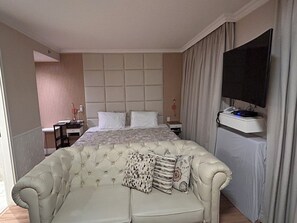 Quarto de hotel com uma cama grande, televisão, escrivaninha com cadeira e sofá.