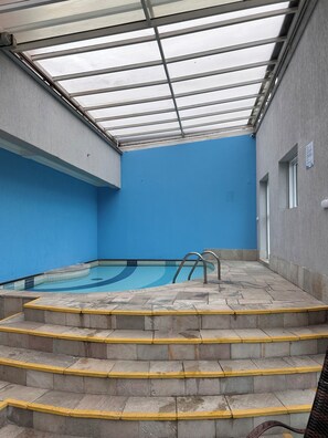 Piscina interna com água cristalina, parede azul e teto de vidro.