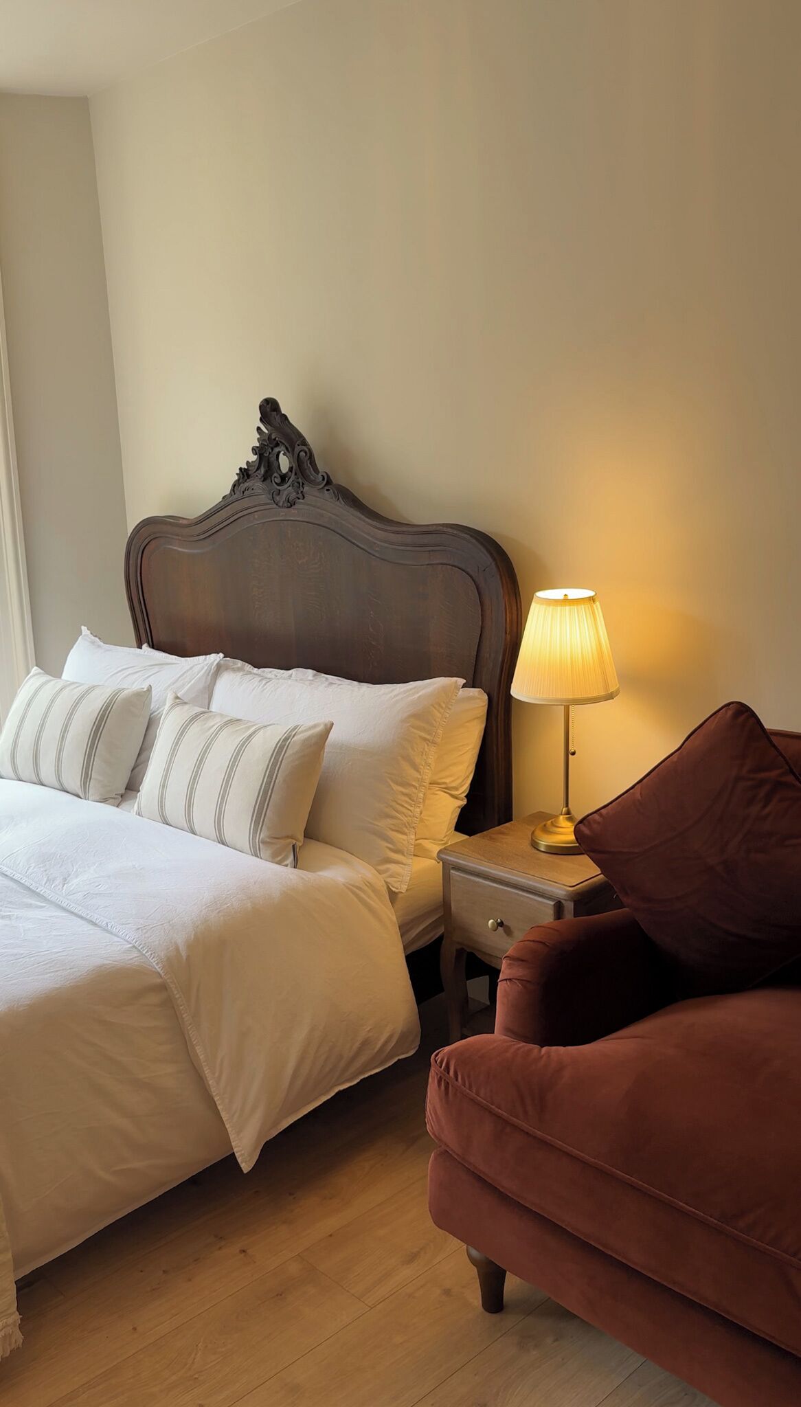A neatly made bed with pillows, a bedside lamp, a wooden nightstand, and a brown armchair in a hotel room.