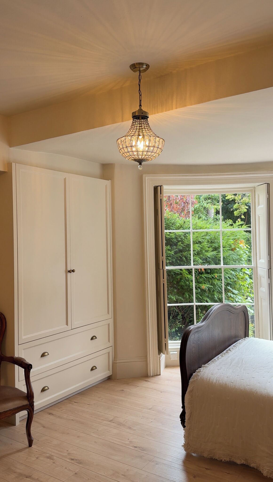 A bedroom with a large wardrobe, a bed, a wooden chair, and a window with a view of greenery.