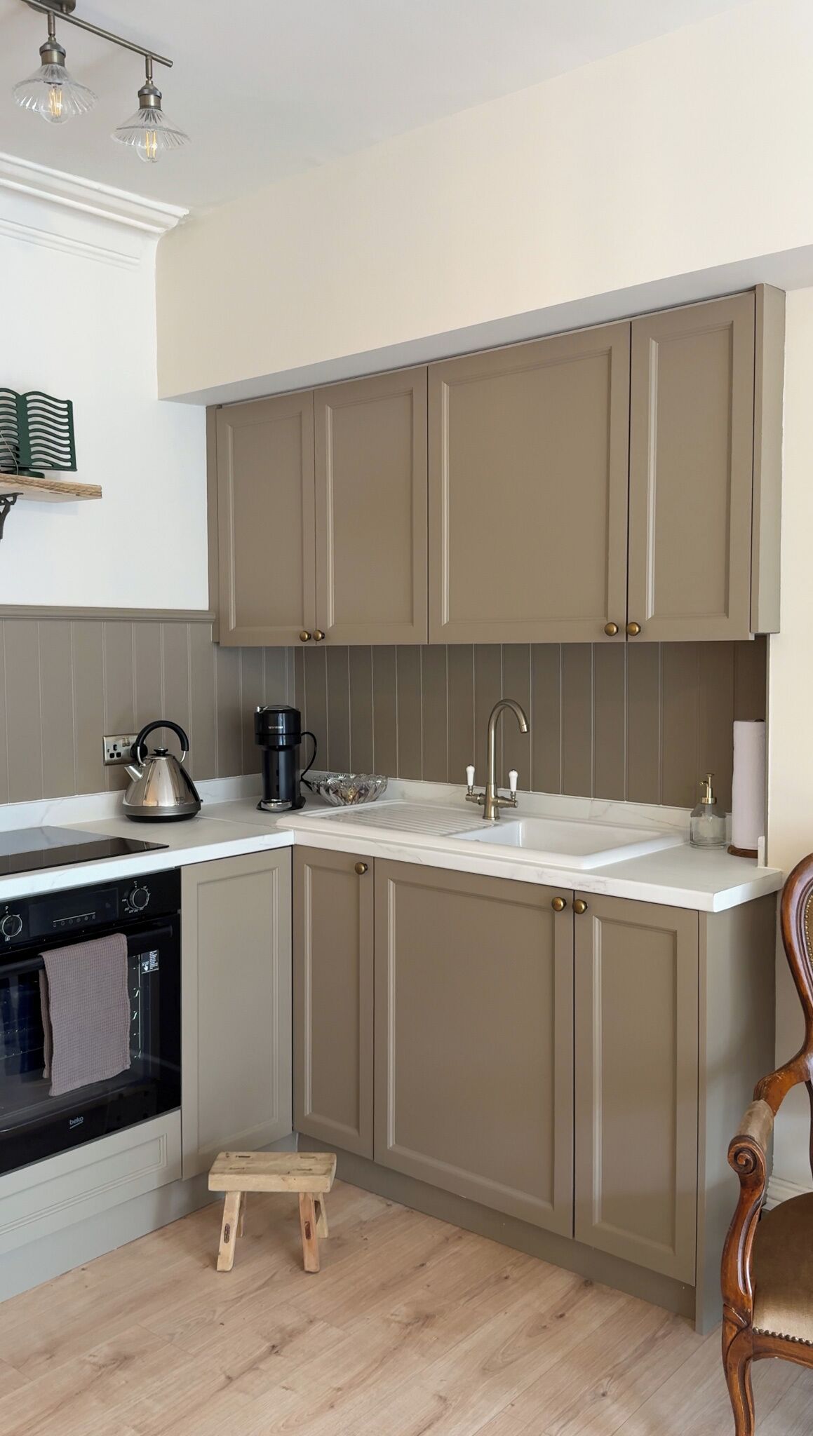 A kitchen with beige cabinets, a white countertop, a kettle, a coffee maker, and a wooden stool.