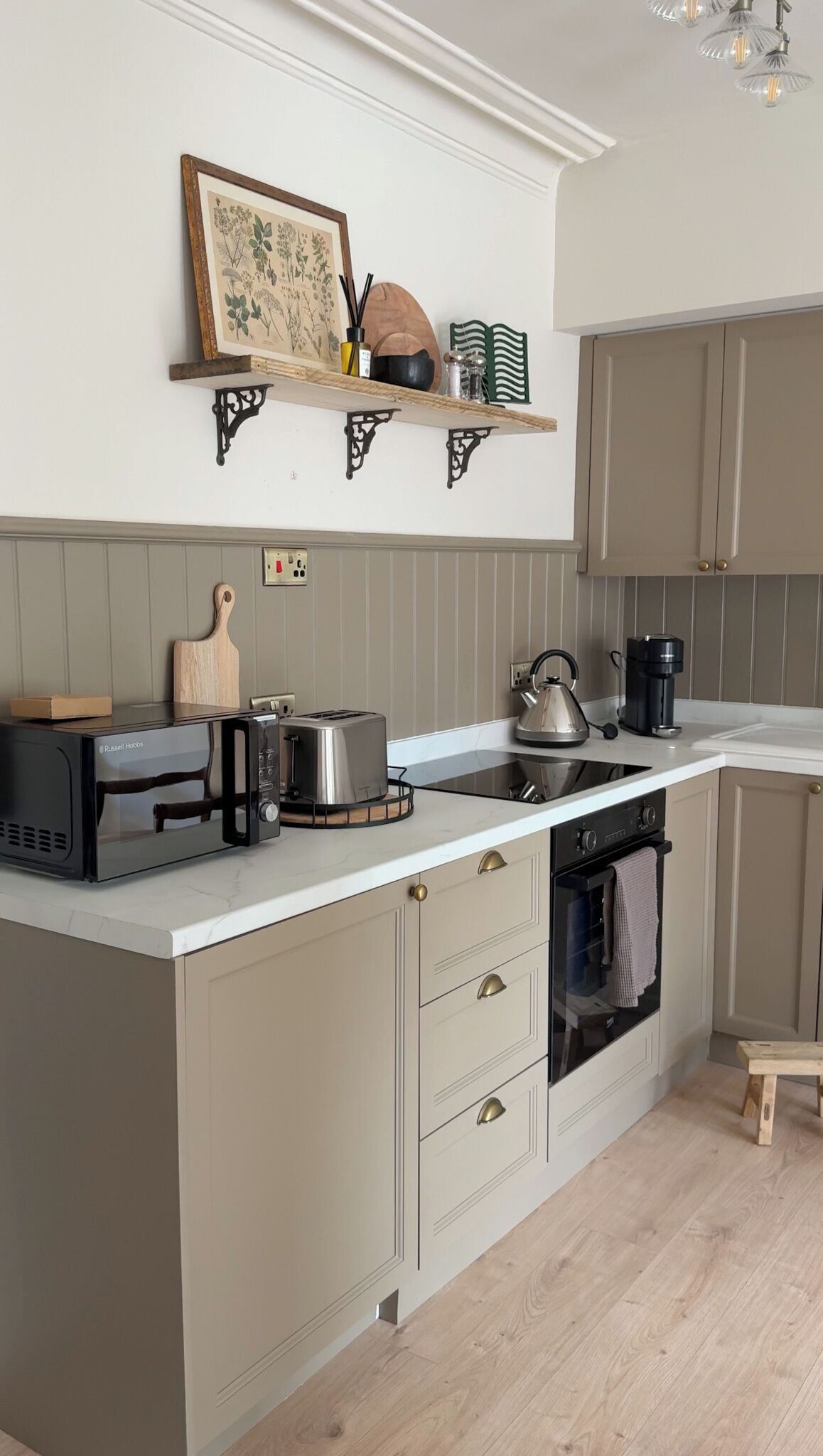 Een keuken met een ingebouwde kookplaat, een waterkoker, een koffiezetapparaat en een broodrooster. Op het aanrecht ligt een houten snijplank en een mes.