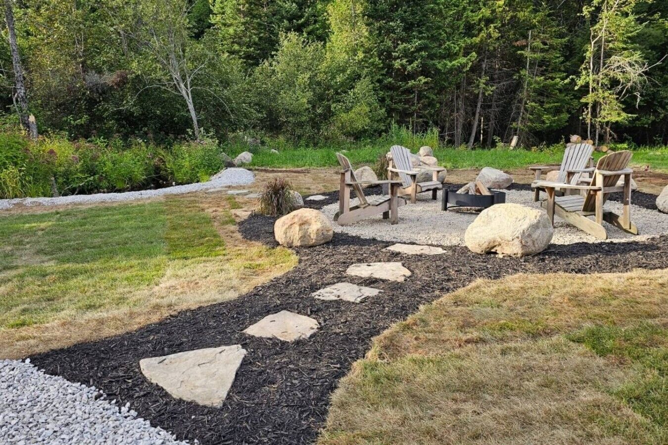 An outdoor seating area with Adirondack chairs and a fire pit, surrounded by a stone pathway and natural landscaping.