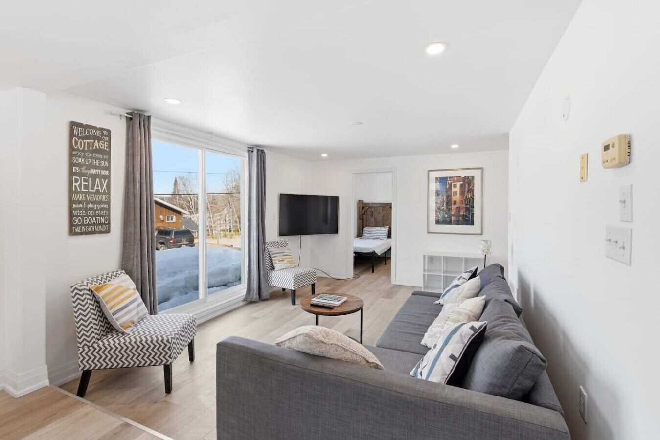 A modern living room with a sofa, armchair, and a coffee table. There is a television mounted on the wall and a painting above it.