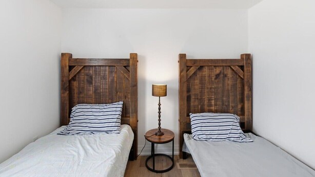 A room with two single beds, each with a wooden headboard and striped pillows. A small round table with a lamp is placed between the beds. - South Baptiste Lake View | Boat Launch | Game Room (Hastings Highlands)