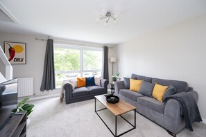 A modern living room with two grey sofas, a wooden coffee table, and a large window with curtains. - 2 Bed Flat Free Parking Next To The Station (West Sussex)
