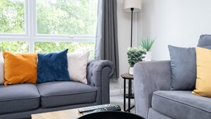 A living room with a grey sofa, a small side table, a lamp, and a potted plant. - 2 Bed Flat Free Parking Next To The Station (West Sussex)