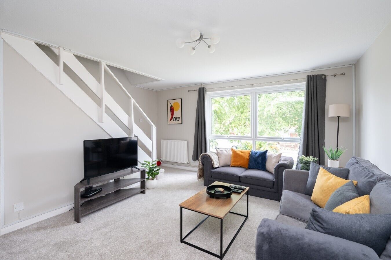 A modern living room with a flat-screen TV, grey sofas with yellow cushions, a wooden coffee table, and a staircase.