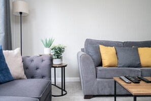 A modern living room with a grey sofa, a smaller grey armchair, a round wooden coffee table, and a side table with a potted plant. - 2 Bed Flat Free Parking Next To The Station (West Sussex)