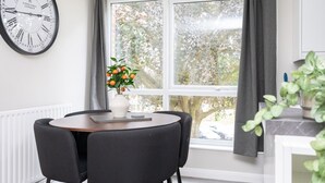 A dining area with a round table, four chairs, a vase with flowers, a clock, and a window with curtains. - 2 Bed Flat Free Parking Next To The Station (West Sussex)