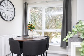 A dining area with a round table, four chairs, a vase with flowers, a clock, and a window with curtains. - 2 Bed Flat Free Parking Next To The Station (West Sussex)