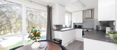 A modern kitchen with white cabinets, a dining table with a vase of fruit, and a view of the outdoors.