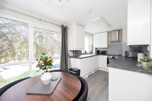 A modern kitchen with white cabinets, a dining table with a vase of fruit, and a view of the outdoors.