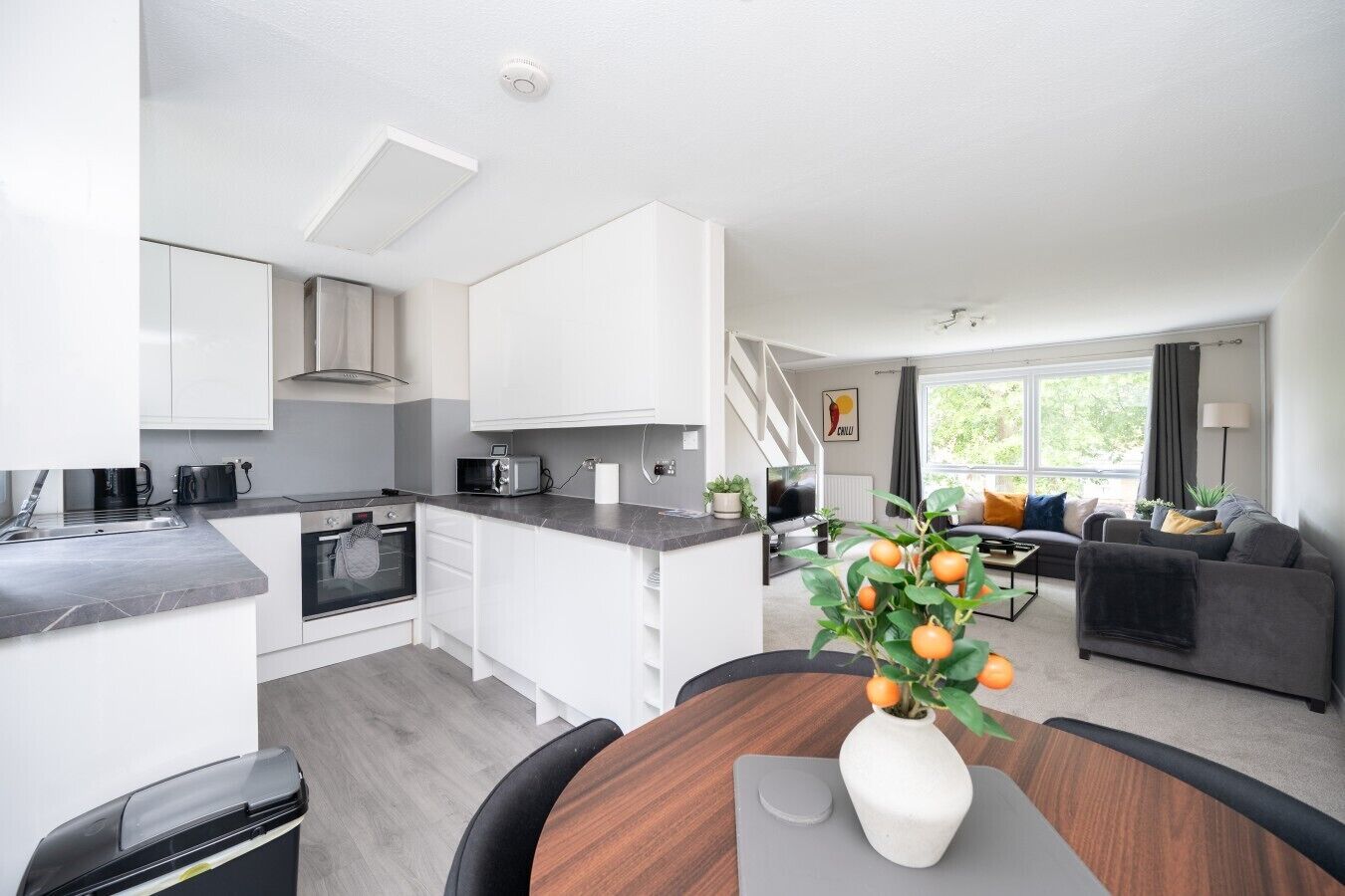 A modern kitchen with white cabinets, a built-in oven, and a microwave. There is a dining area with a wooden table and chairs, and a living room with a sofa and a TV.