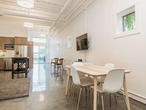A modern kitchen with stainless steel appliances, a dining area with a long table and chairs, and a television mounted on the wall. - Modern Loft Haven | BBQ, Sleeps 6 | (Calgary)