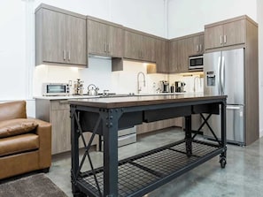 A modern kitchen with wooden cabinets, a stainless steel refrigerator, and a central island with a metal table. - Modern Loft Haven | BBQ, Sleeps 6 | (Calgary)