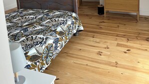 A bedroom with a sloped ceiling, a bed with a patterned bedspread, a bedside table with a lamp, a wooden dresser, and a round mirror on the wall. - Farm cottage (Le Haut-Corlay)