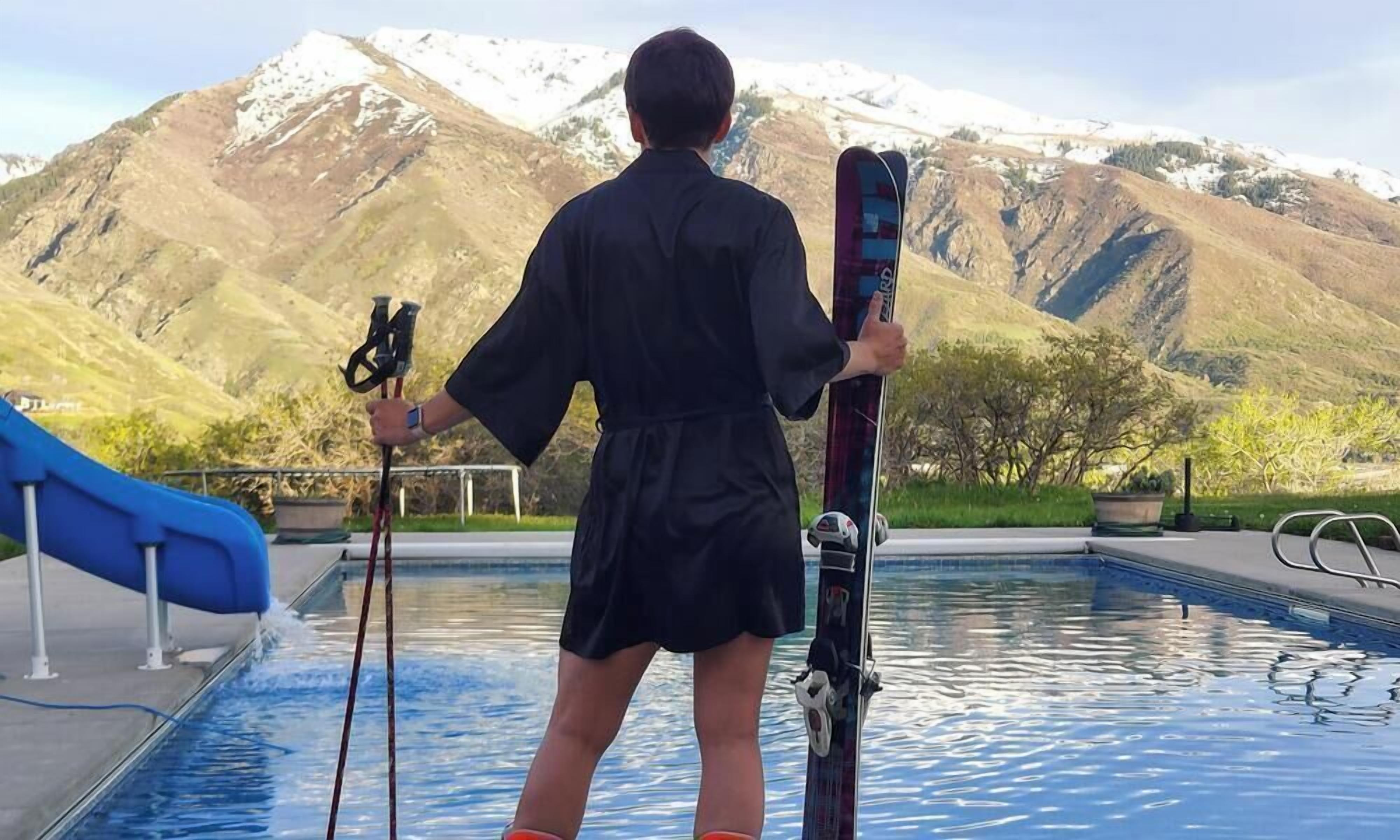 Une personne tenant des skis près d’une piscine, avec des montagnes en arrière-plan.