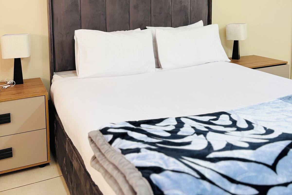 Habitación de hotel con una cama, dos mesitas de noche, un aparador y una manta decorativa estampada.