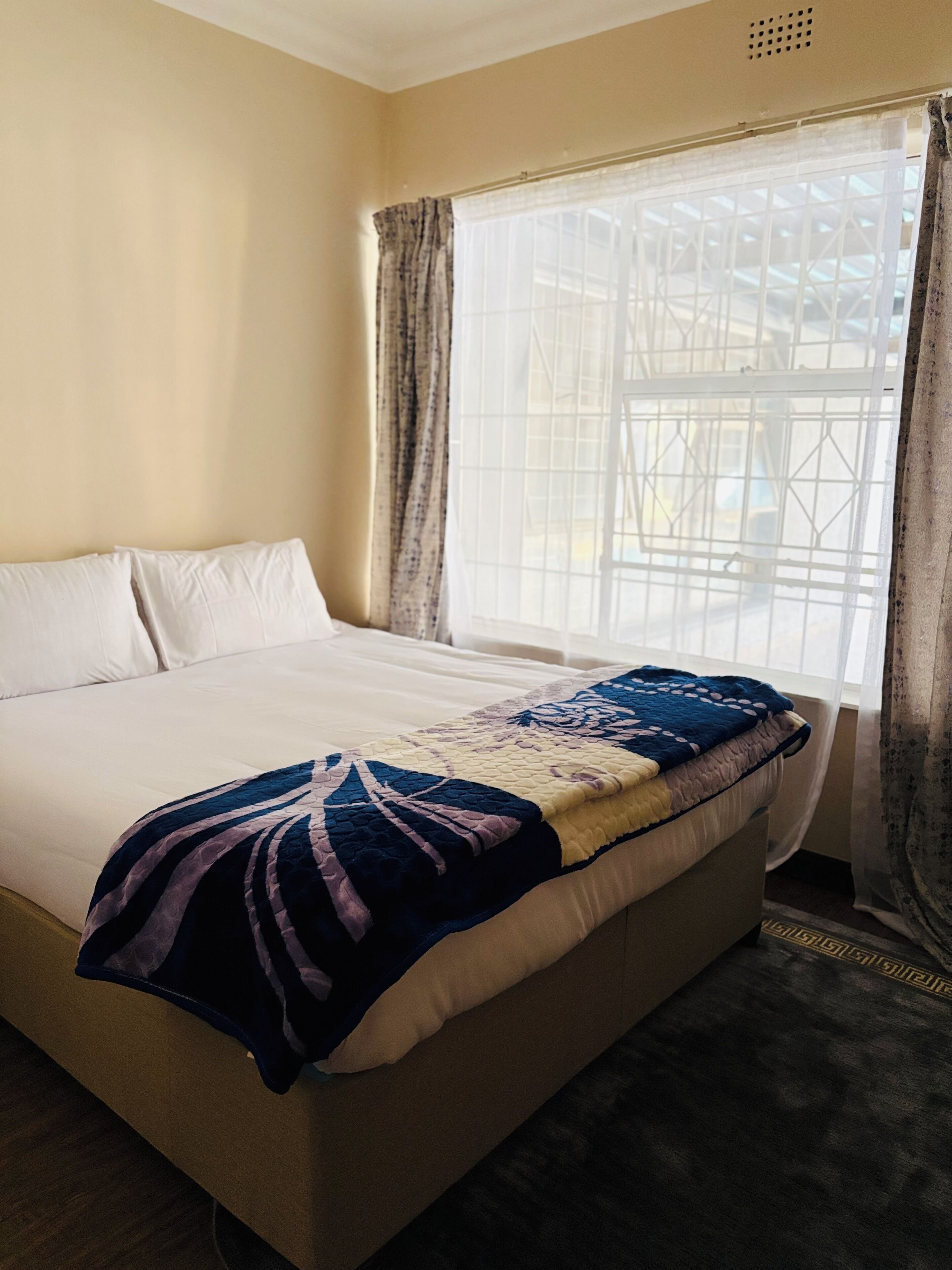 Una cama con un edredón estampado, una ventana con vistas a una edificación y cortinas.