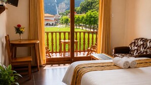 Habitación de hotel con cama, escritorio con silla, televisor montado en la pared y balcón con vistas a una cascada y vegetación.