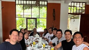 Un grupo de personas sentadas alrededor de una mesa con alimentos para el desayuno, en una habitación con ventanas y un ventilador de techo.