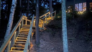 A wooden staircase leading to a building, surrounded by trees, with two blue Adirondack chairs on a wooden deck. - NEW Luxury Riverfront Home, large deck, river tubing, kayaks, 5 min to Downtown (Hayward)