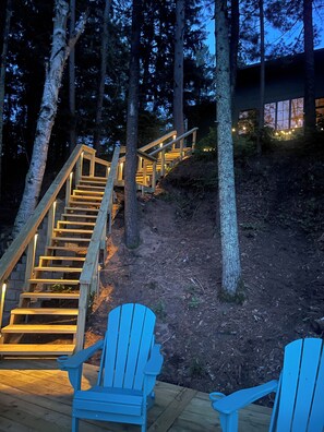 Een houten trap die naar een gebouw leidt, omringd door bomen, met twee blauwe Adirondack-stoelen op een houten terras.