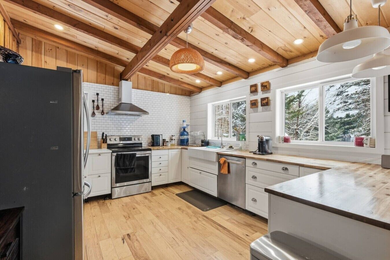A modern kitchen with wooden beams, white cabinets, a stainless steel oven, and a large window providing a view of trees.