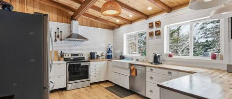 A modern kitchen with wooden beams, white cabinets, a stainless steel oven, and a large window providing a view of trees.