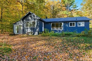 Une cabane dotée d’une grande fenêtre et d’une porte, entourée d’une forêt aux feuilles d’automne.