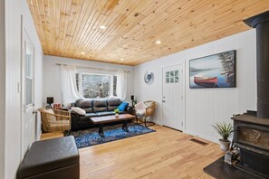 A cozy living room with a wooden ceiling, a black leather sofa, a blue patterned rug, a wicker chair, and a fireplace. - Serene Otter Lake Waterfront, Dock + Fire Pit (Huntsville)