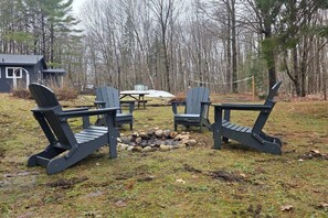 Un espace de détente circulaire, composé de fauteuils Adirondack disposés autour d’un foyer, situé dans une zone boisée avec une maison en arrière-plan.