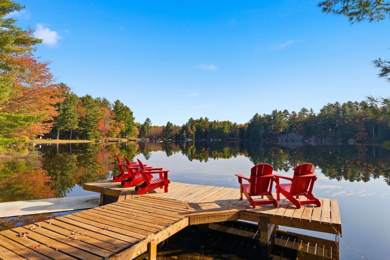 A wooden dock with red chairs extending over a calm lake, surrounded by trees with autumn foliage.