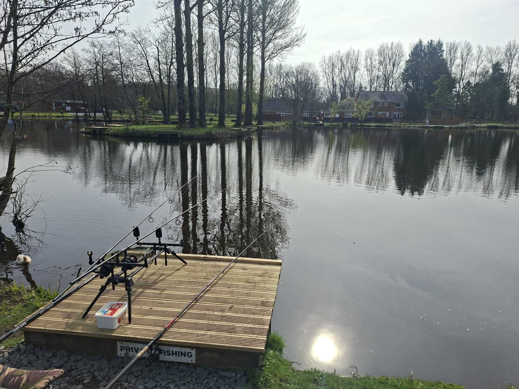 A fishing platform by a calm lake with a wooden deck, surrounded by trees and houses in the background.