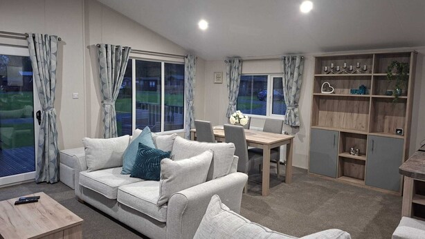 A modern living room with a sofa, a dining table, and a built-in bookshelf. - Holiday Lodge with fishing set in tranquil surroundings (Forton)