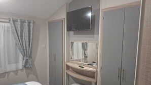 A bedroom with a bed, a TV mounted above a mirror, a wardrobe, and a small stool. - Holiday Lodge with fishing set in tranquil surroundings (Forton)