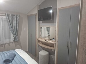 A bedroom with a bed, a TV mounted above a mirror, a wardrobe, and a small stool. - Holiday Lodge with fishing set in tranquil surroundings (Forton)