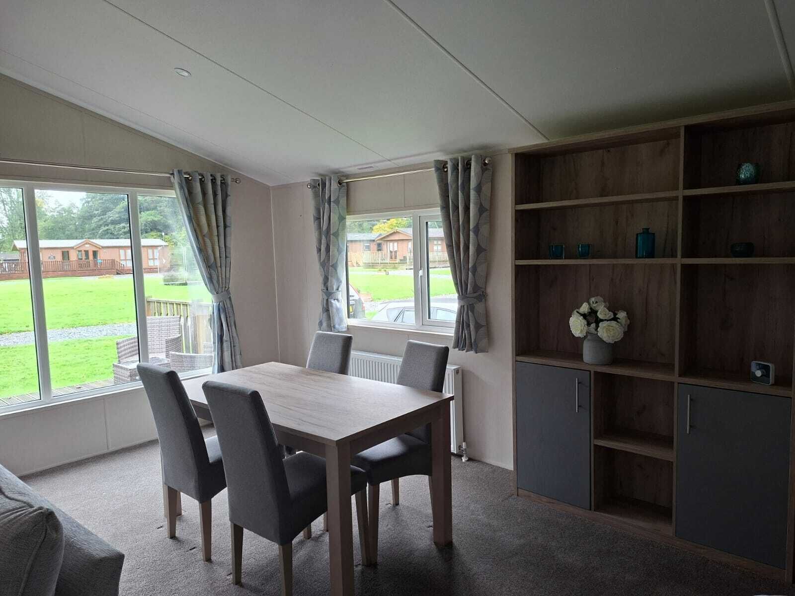 A dining area with a table and chairs, large windows with curtains, and a built-in shelving unit with decorative items.