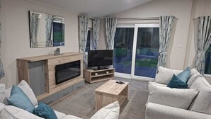 A living room with a TV, wooden cabinet, sofa, and a sliding glass door. - Holiday Lodge with fishing set in tranquil surroundings (Forton)