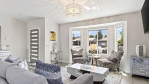 Una sala de estar moderna con un sofá, una mesa de centro y sillones estampados. Se observa una araña, un televisor y vistas al exterior.