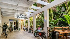 Et overdækket udendørs opholdsområde med borde og stole, et skilt der siger 'KAVA GARDEN', og en motorcykel parkeret i nærheden.