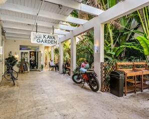 Ein überdachter Außenbereich mit Tischen und Stühlen, ein Schild mit der Aufschrift 'KAVA GARDEN' und ein geparktes Motorrad in der Nähe.