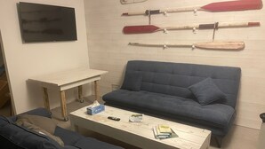 A living room with a blue sofa, a white coffee table with books and tissues, a small table with a remote, and a wall-mounted display of paddles. - La maison des plages sleeps 14 (Erquy)