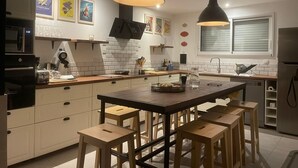 A modern kitchen with a central island, wooden stools, pendant lighting, and a flat-screen TV mounted on the wall. - La maison des plages sleeps 14 (Erquy)