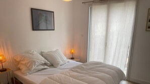 A bedroom with a bed, two bedside lamps, a window with curtains, and a framed picture on the wall. - La maison des plages sleeps 14 (Erquy)