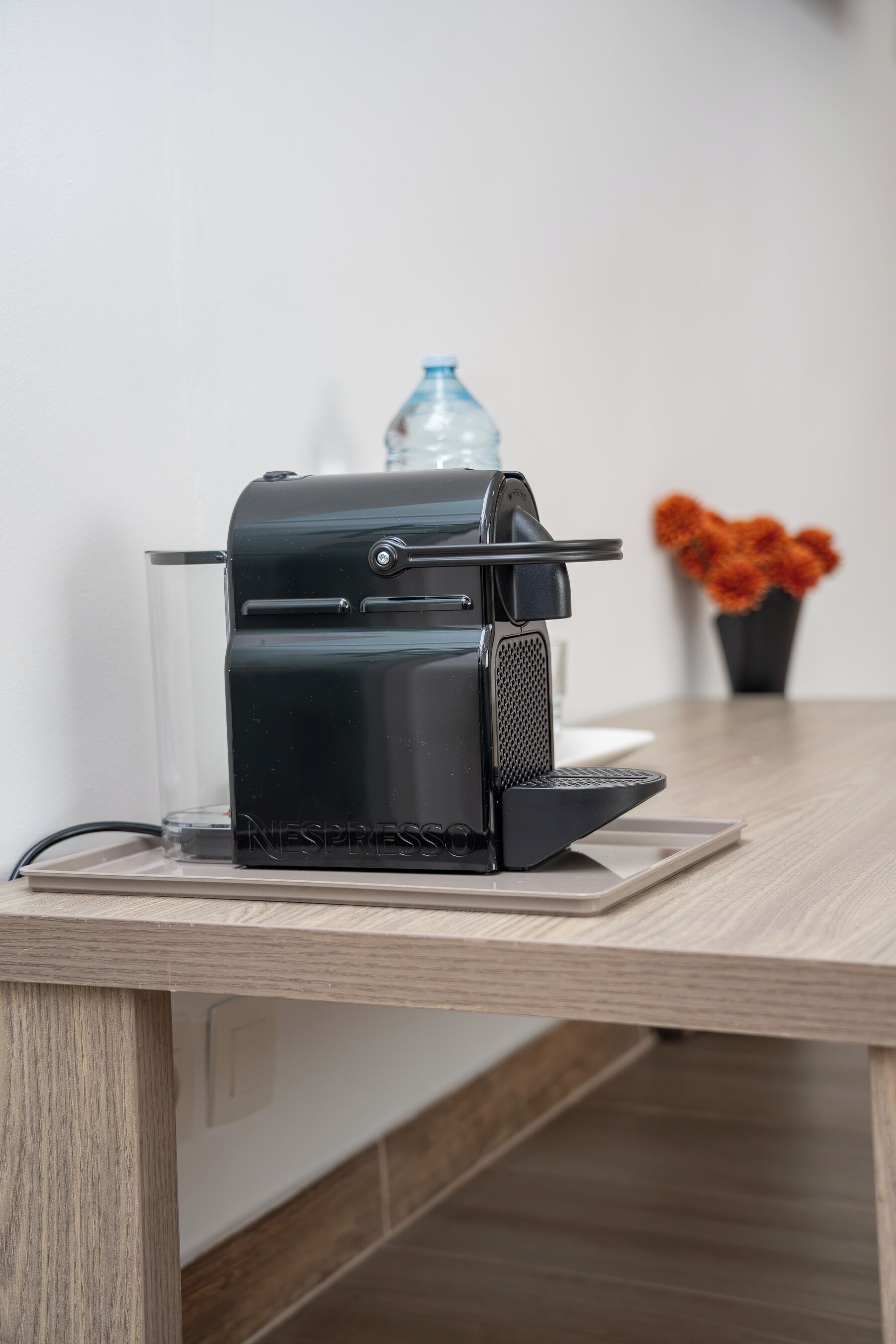 A Nespresso coffee machine on a wooden table with a clear glass, a water bottle, and a potted plant in the background.