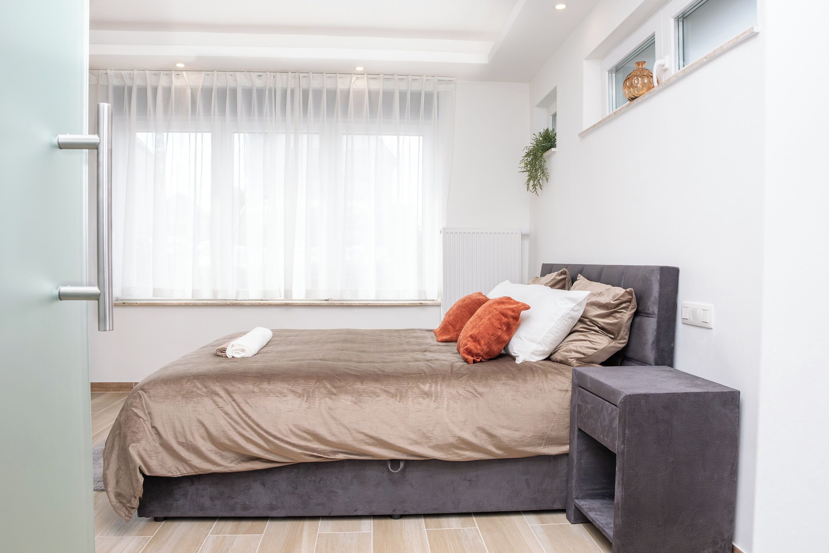 Un dormitorio con cama, almohadas, una mesita de noche, una ventana con cortinas, un radiador y una planta en la pared.