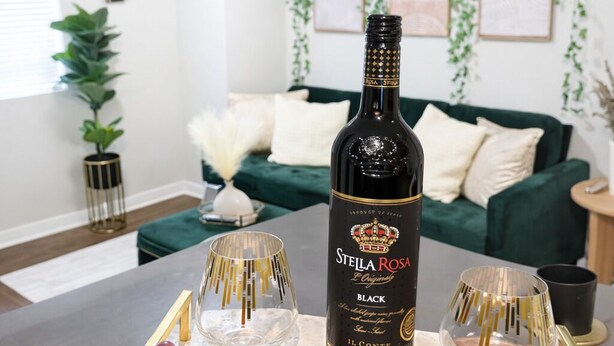 A bottle of Stella Rosa Black wine on a tray with grapes and glasses in a modern living room with a green couch and decorative plants.